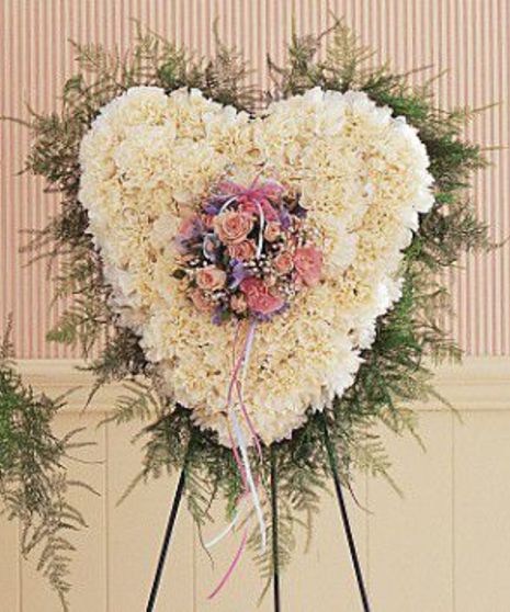 A heart-shaped floral arrangement of white flowers with a central bouquet of pink and purple flowers on a stand, placed against a striped wallpaper background