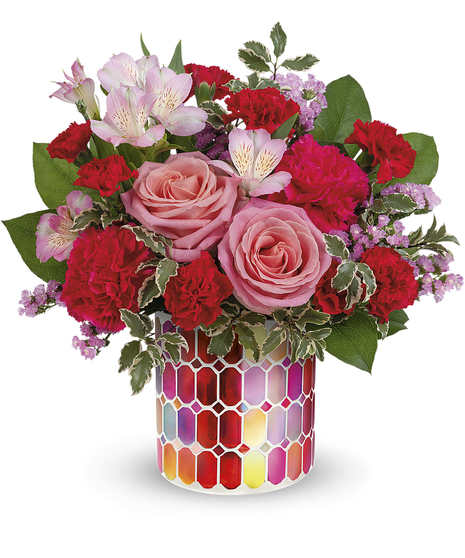 Bouquet of pink roses, red carnations, and assorted flowers arranged in a colorful, hexagon-patterned vase; green leaves surround the flowers, providing contrast.