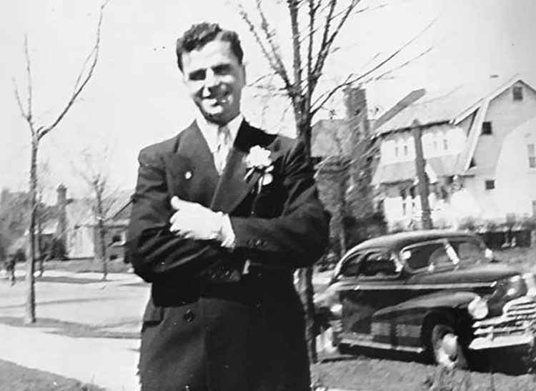 Pictured as a young man, Frank Mancuso Sr. poses in a suit and tie, beside a classic car